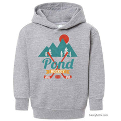 A heather gray toddler pullover hoodie featuring a graphic design on the chest with mountains, pine trees, a sun, crossed hockey sticks, and the text 'Pond Hockey'.