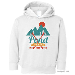 A white toddler pullover hoodie featuring a graphic design on the chest with mountains, pine trees, a sun, crossed hockey sticks, and the text 'Pond Hockey'.